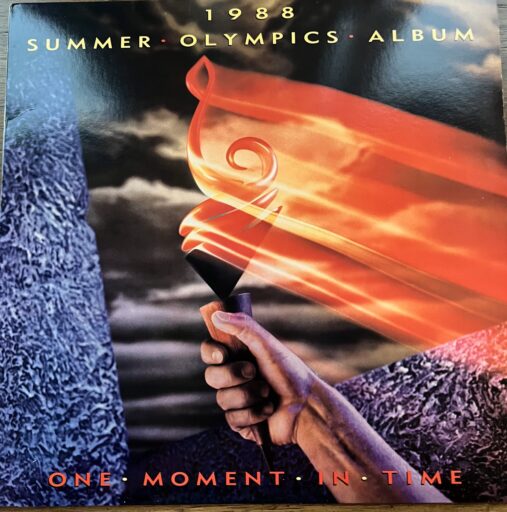 A cover of a "1988 Summer Olympics Album" titled "One Moment in Time." It features a close-up of a hand holding the Olympic torch, with the flame burning brightly and swirling upwards. The background is dark with a hint of clouds, emphasizing the glowing torch. The text and imagery are set against a black backdrop.