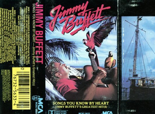 A cassette tape case. The cassette is titled "Jimmy Buffett - Songs You Know By Heart, Jimmy Buffett's Greatest Hit(s)". The cover art features Jimmy Buffett playing a guitar, with palm trees and a pink and blue sky in the background, suggesting a tropical setting.